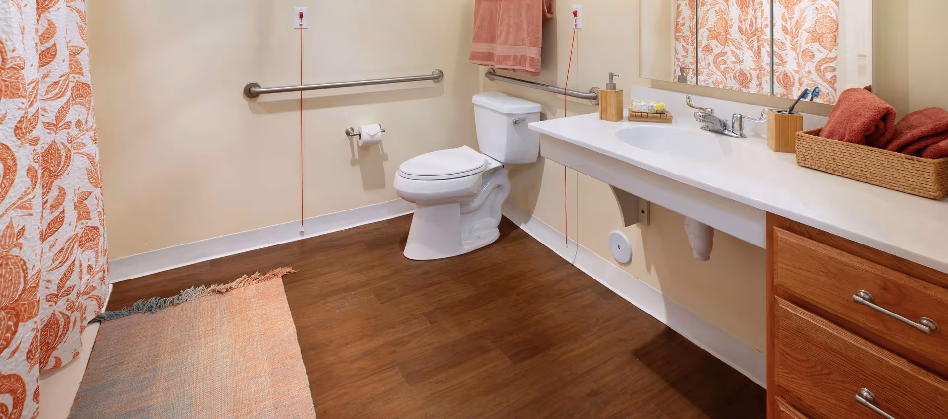 A bathroom with a white toilet, a long white sink countertop with a faucet, a large mirror above the sink, and wooden cabinets below. There are grab bars on the walls near the toilet and sink. A patterned orange and white shower curtain is partially visible on the left side, along with a small rug on the floor. Towels and toiletries are neatly arranged on the countertop.