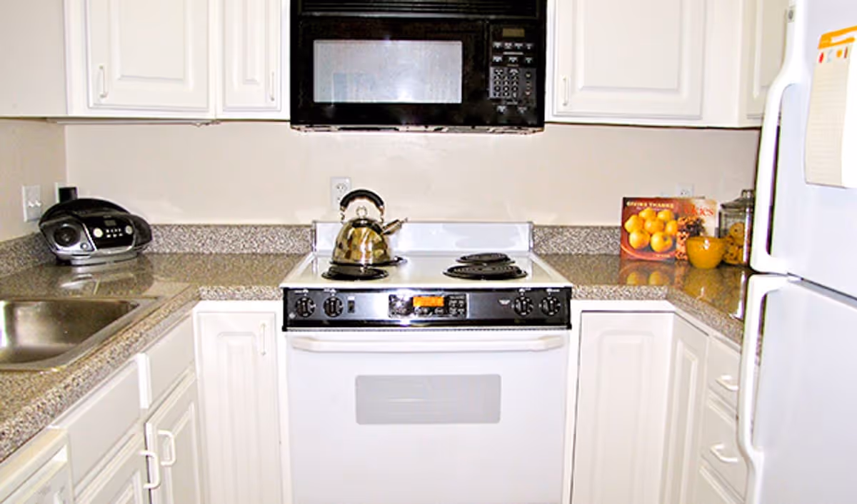 A compact kitchen with white cabinets and granite countertops. There is a stainless steel sink on the left, a white electric stove with a kettle on top in the center, and a white refrigerator on the right. Above the stove is a black microwave. On the countertop to the right, there is a bowl of oranges and some kitchen items.