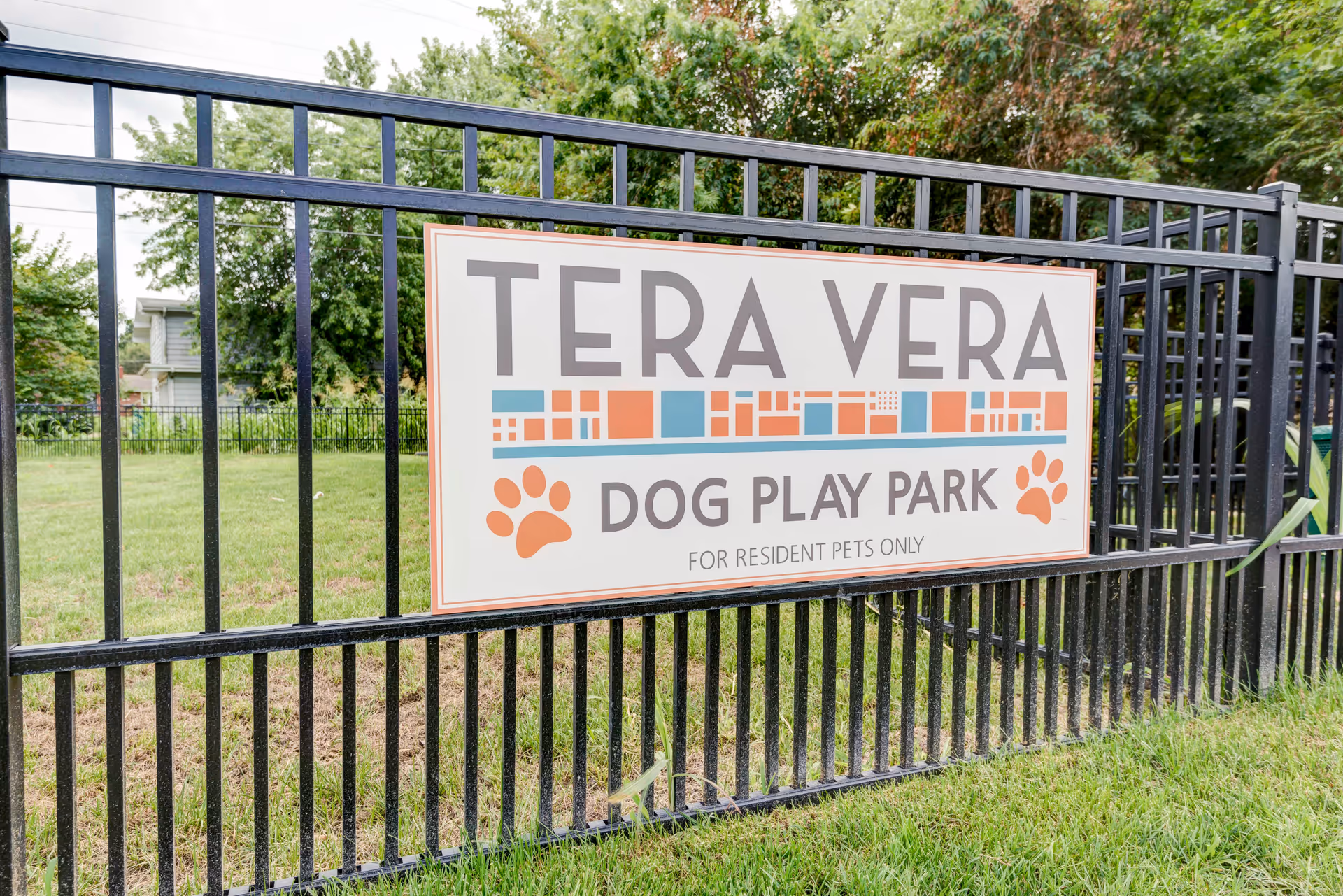 A sign on a black metal fence that reads "TERA VERA DOG PLAY PARK" with grass and trees in the background.