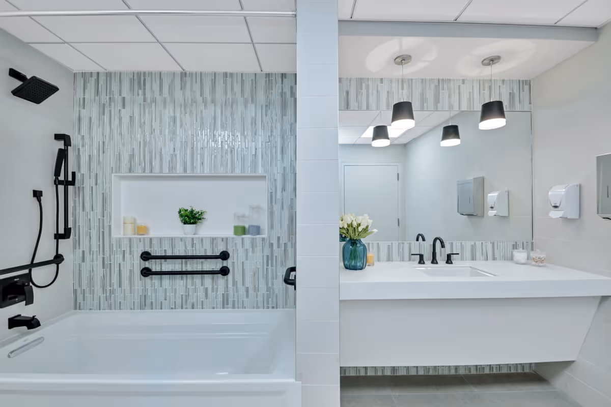 Modern bathroom with a white bathtub featuring black fixtures and a built-in niche with candles and a small plant. The wall behind the tub is decorated with vertical gray and white tiles. Adjacent to the tub is a white vanity with a large mirror, black faucet, and a vase with white flowers. The bathroom has a clean, contemporary design with light-colored walls and ceiling tiles.