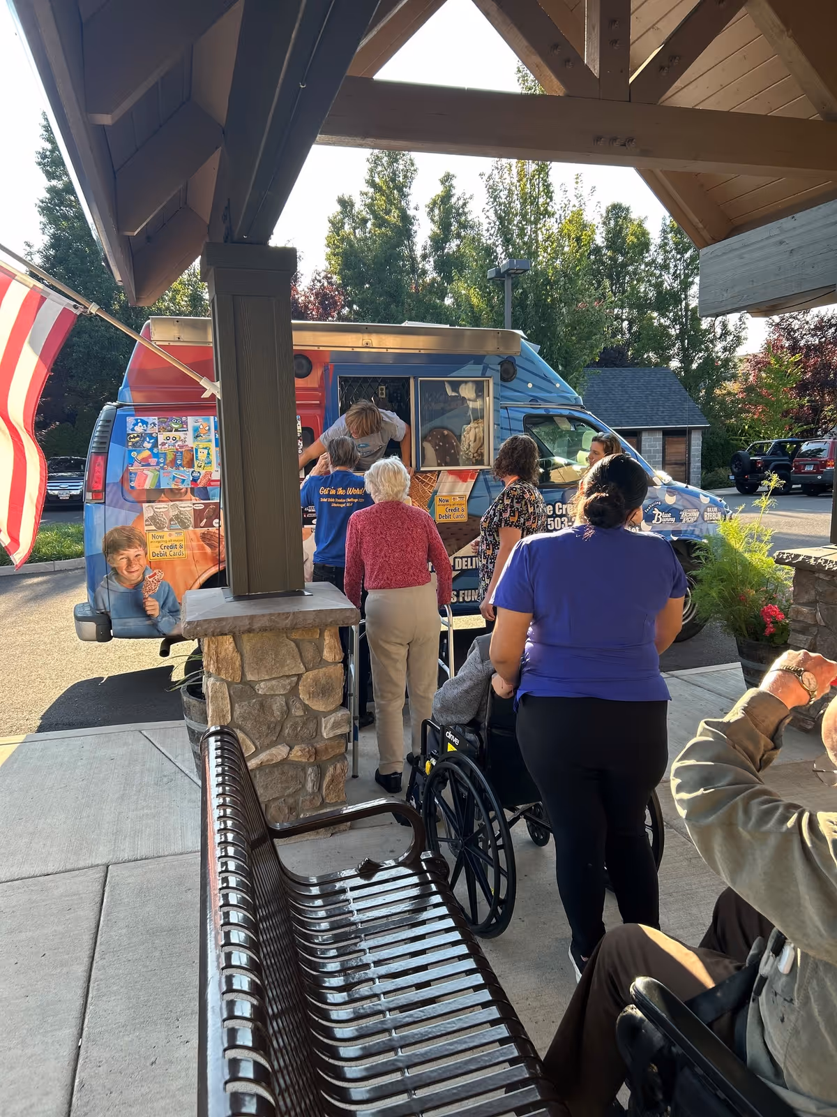 A group of people, including elderly individuals and caregivers, are gathered outside under a covered area near a stone pillar, waiting in line at an ice cream truck. The truck is decorated with colorful images of ice cream treats. There is an American flag on the left side of the image and trees in the background.