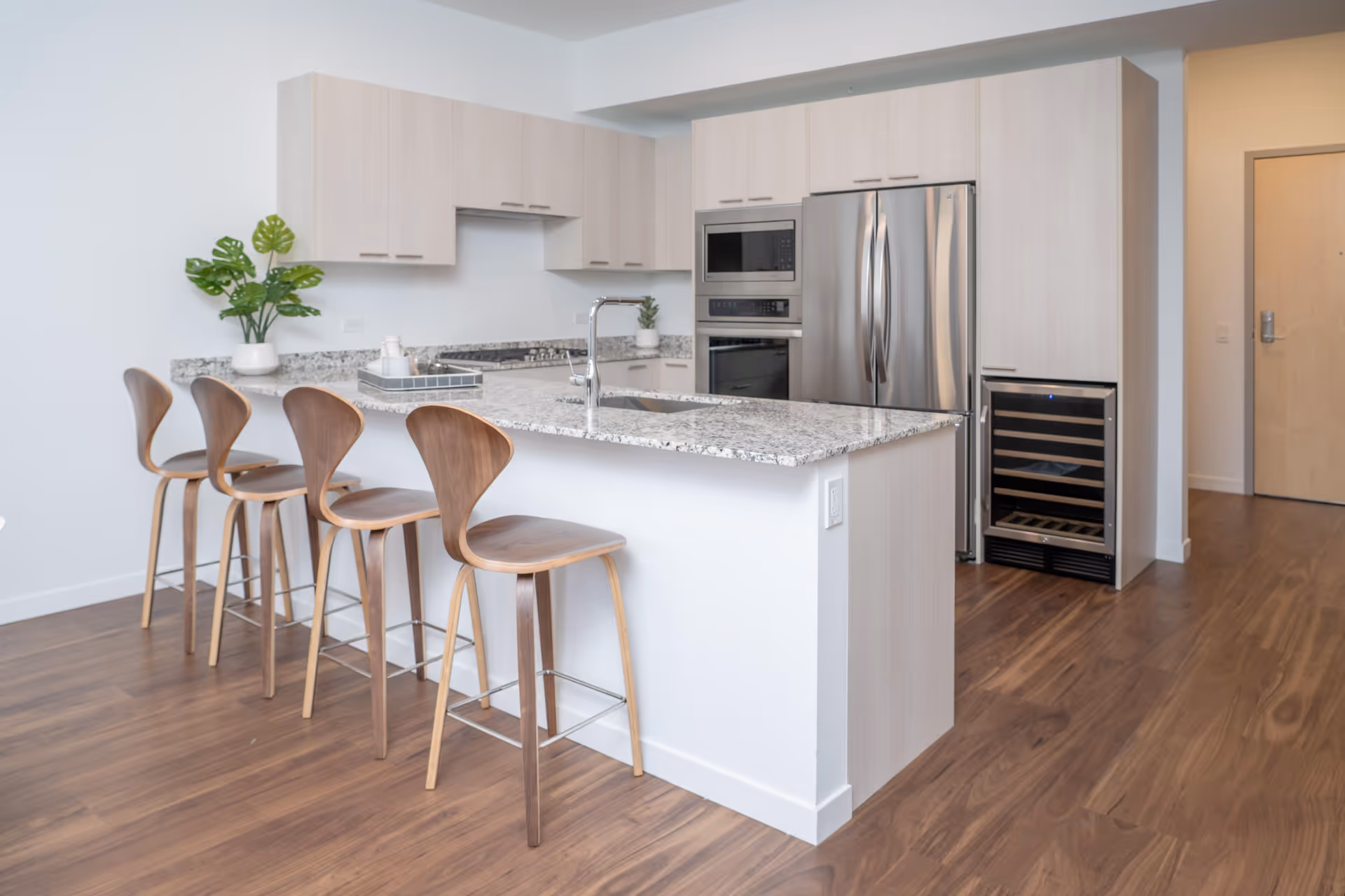 Modern kitchen with a granite island, four wooden bar stools, stainless steel appliances, and light wood cabinetry.