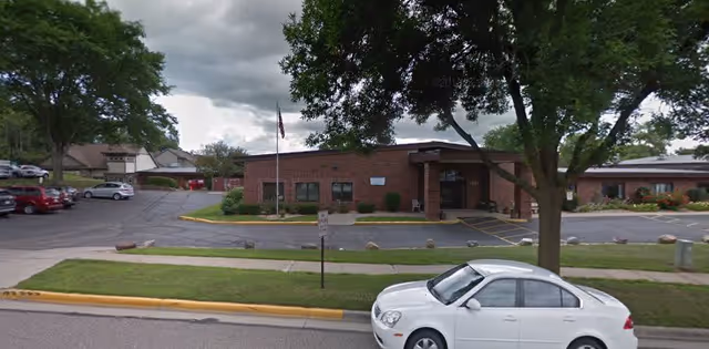 Front exterior view of a single-story brick building with a covered entrance, surrounded by a parking lot with several parked cars and a large tree in the foreground. The sky is cloudy.