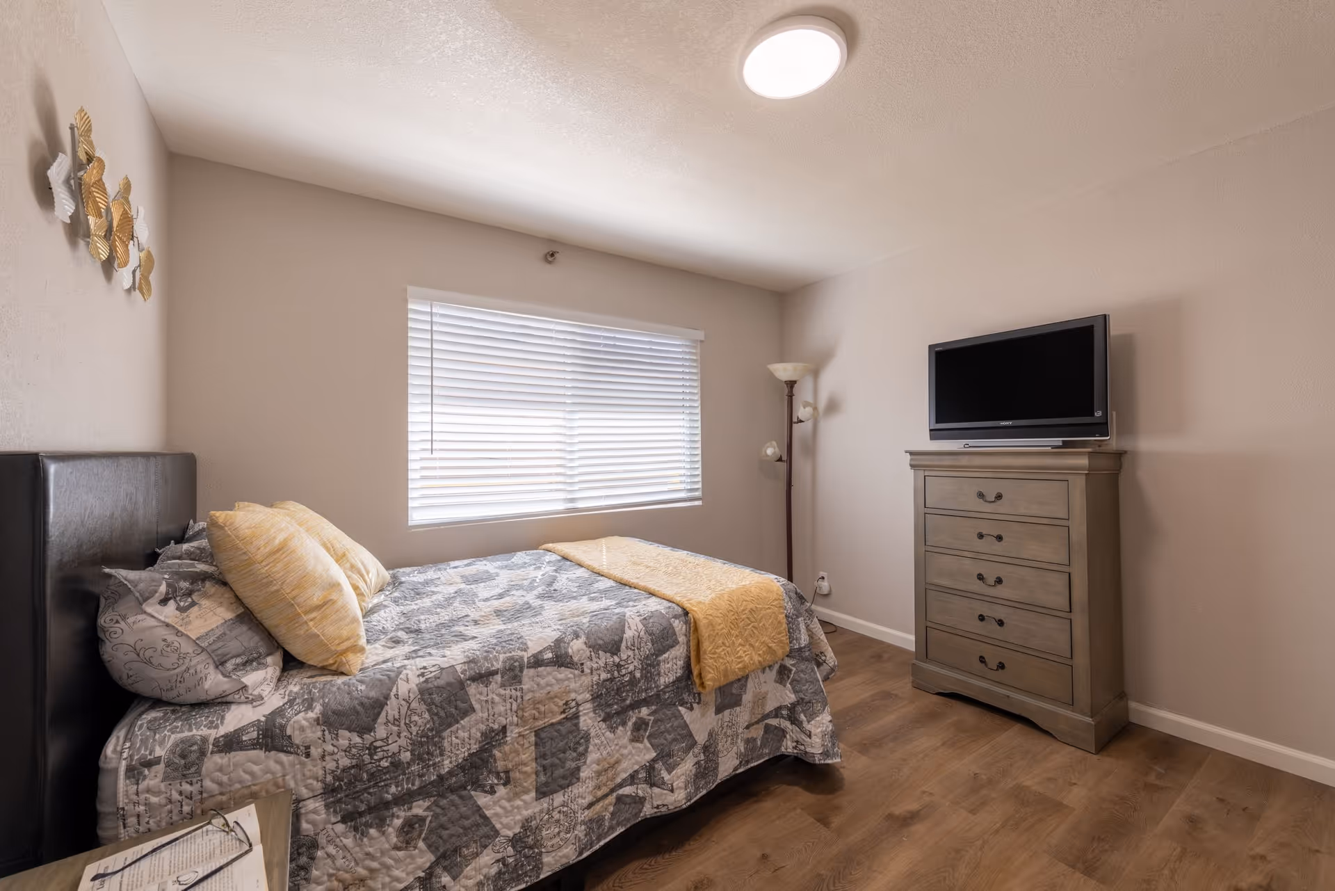 A bedroom with a bed covered in a patterned quilt and yellow pillows, a window with blinds, a wooden dresser with a flat-screen TV on top, a floor lamp in the corner, and light-colored walls and flooring.