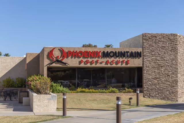Exterior front of the Phoenix Mountain Post-Acute building showing the facility sign above glass windows and a landscaped entrance.
