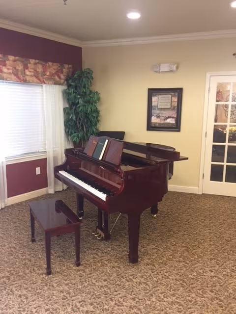 A polished grand piano with a matching bench in a carpeted communal room with a potted plant, framed art, and French doors.