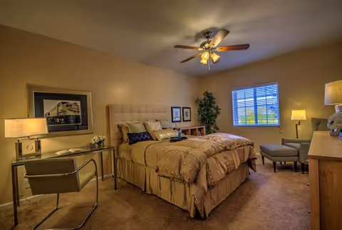 A warmly lit bedroom with a made bed and headboard, desk and chair, armchair, lamps, a ceiling fan, and a window.