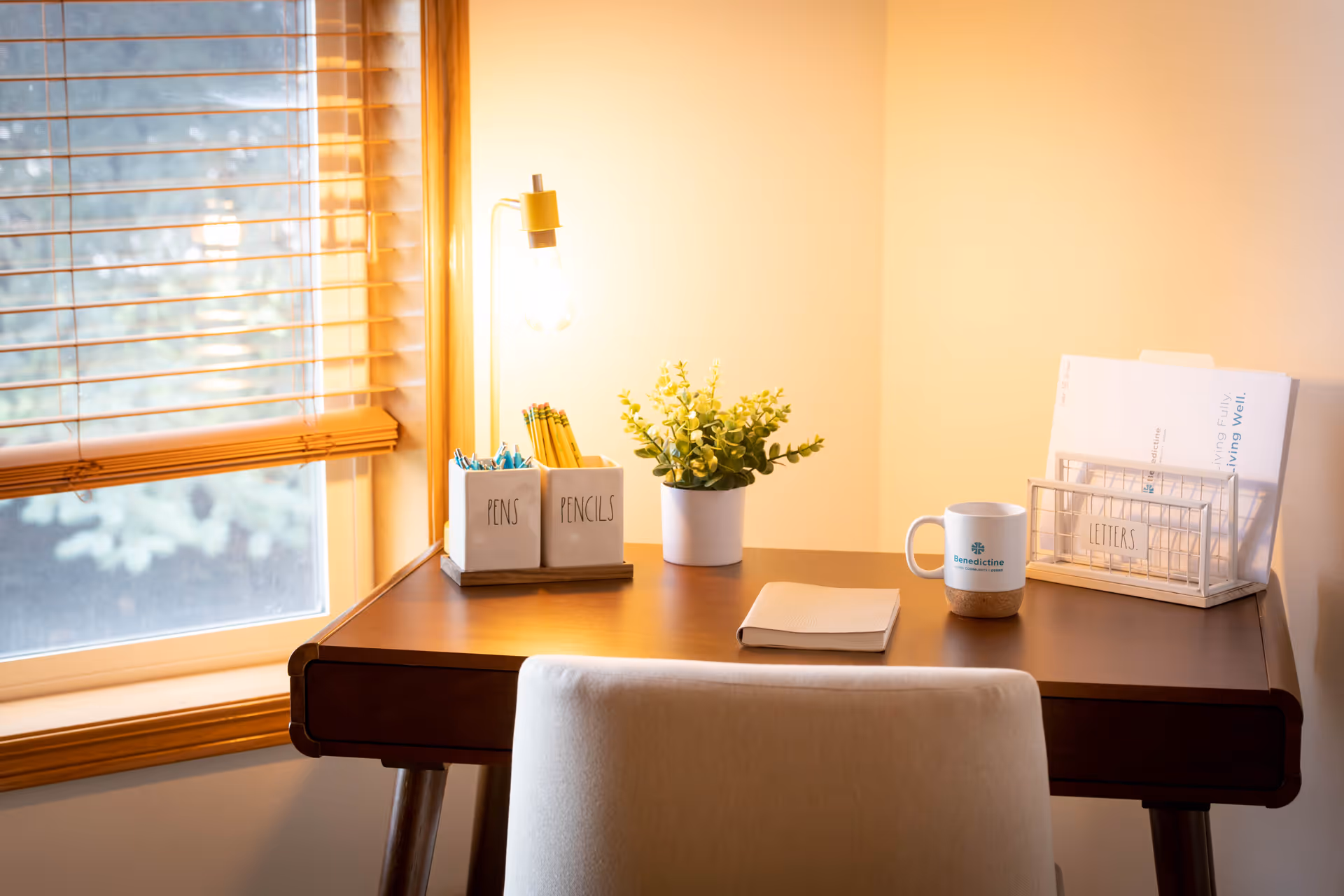 A cozy desk setup near a window with wooden blinds. On the desk are containers labeled 'PENS' and 'PENCILS' holding writing utensils, a small potted plant, a closed notebook, a white mug with the Benedictine logo, and a wire organizer holding papers and letters. A warm light bulb hangs above the desk, creating a comfortable ambiance.