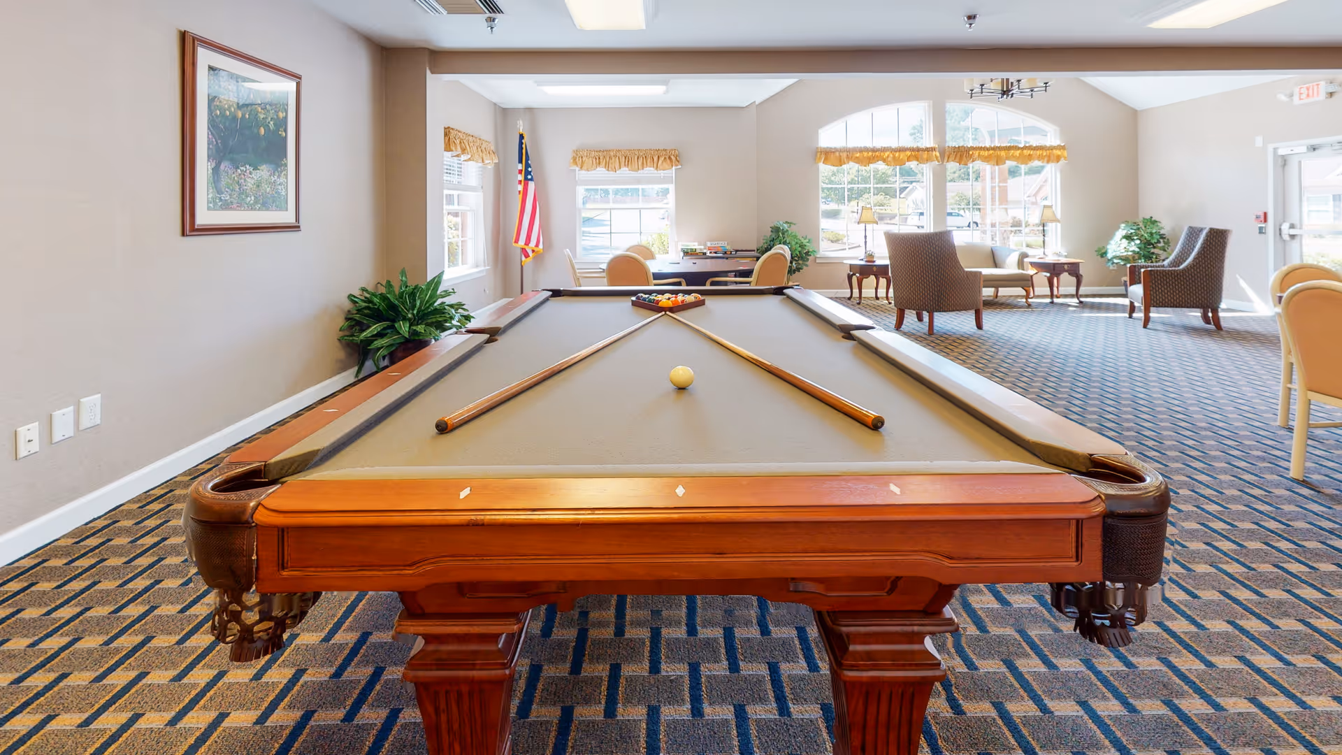 Long wooden pool table centered in a bright communal lounge with chairs, large windows, plants, and an American flag.