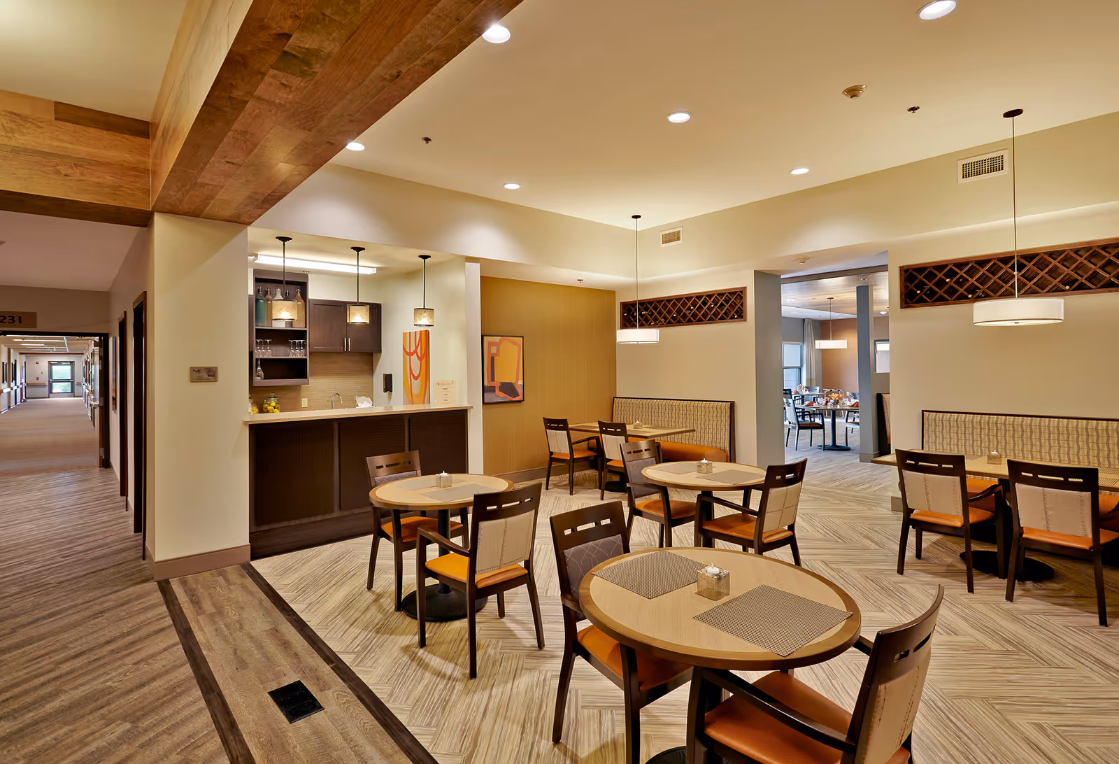 A well-lit dining area in a senior living facility with round tables and chairs arranged neatly. The room features a small bar or serving counter with pendant lights hanging above it. There are booth-style seating areas along the walls, decorative wall art, and a hallway leading to other parts of the facility.