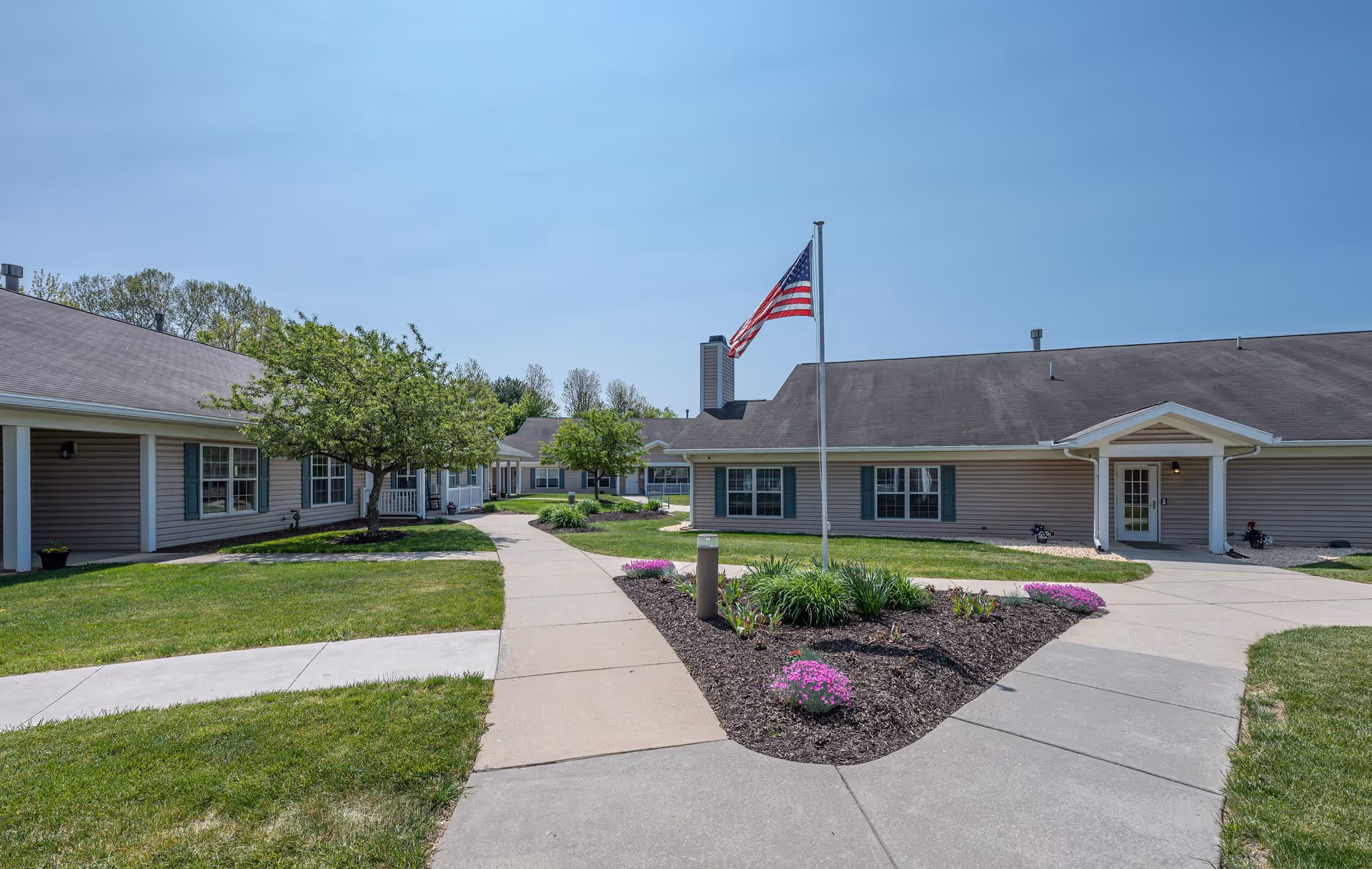 Exterior view of a single-story senior living community building with beige siding and a gray roof. A concrete walkway leads through a landscaped area with green grass, small trees, and flower beds. An American flag is flying on a flagpole near the center of the image under a clear blue sky.