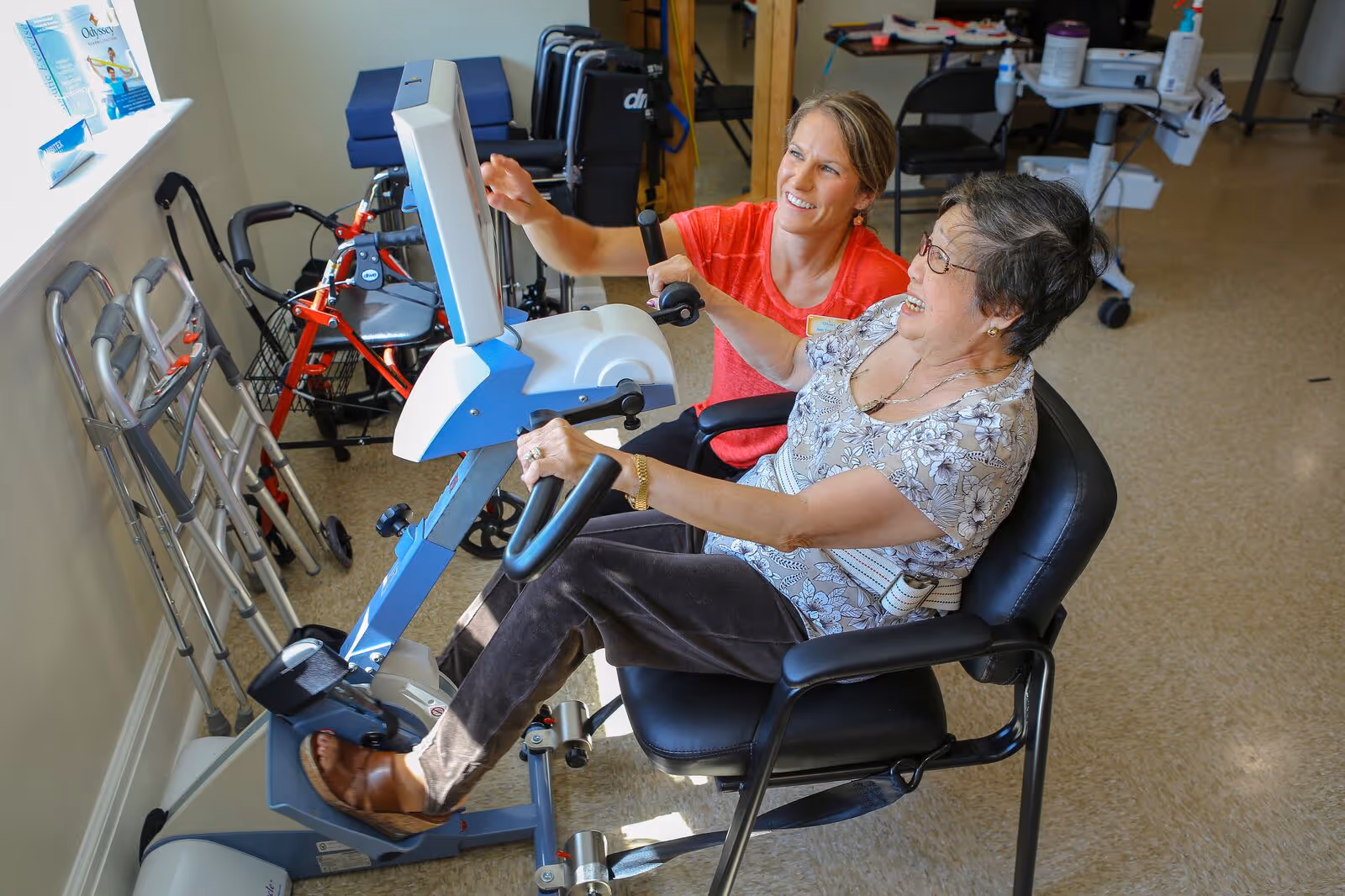 An elderly woman seated on a black chair is using a recumbent exercise bike with the assistance of a smiling caregiver in a red shirt. The room has medical equipment such as walkers and wheelchairs lined up against the wall.
