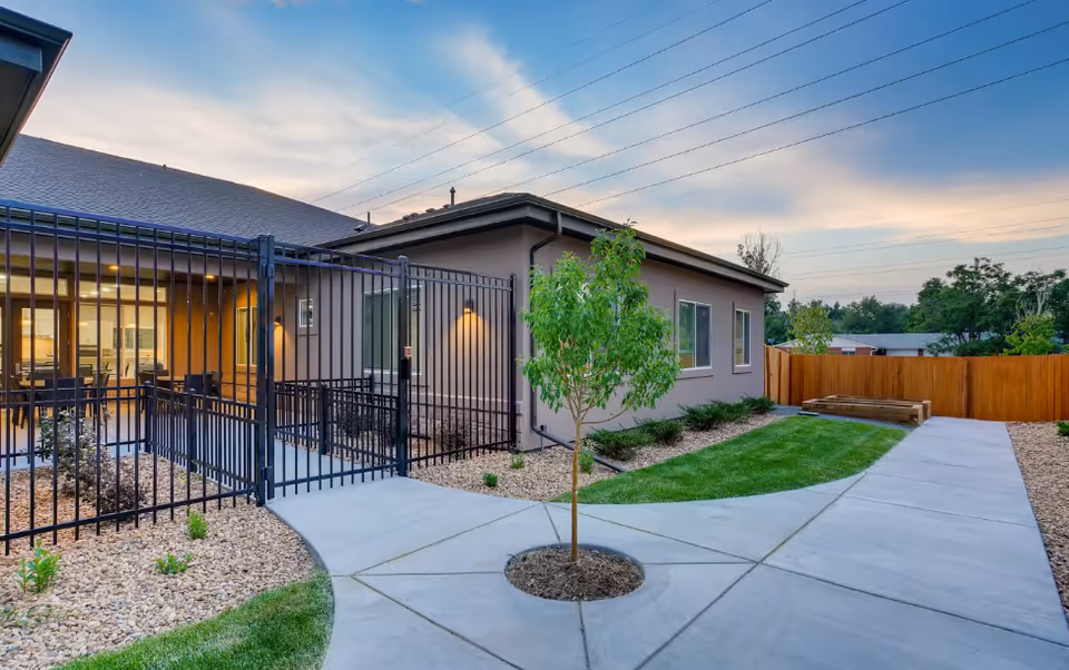 Courtyard of an assisted living building showing a young tree, paved paths, a fenced patio and the building exterior at dusk.