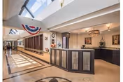 Interior view of a nursing center reception area with a front desk, decorative lighting, and skylights allowing natural light to fill the space. The hallway extends to the left with windows and patriotic bunting hanging near the ceiling.
