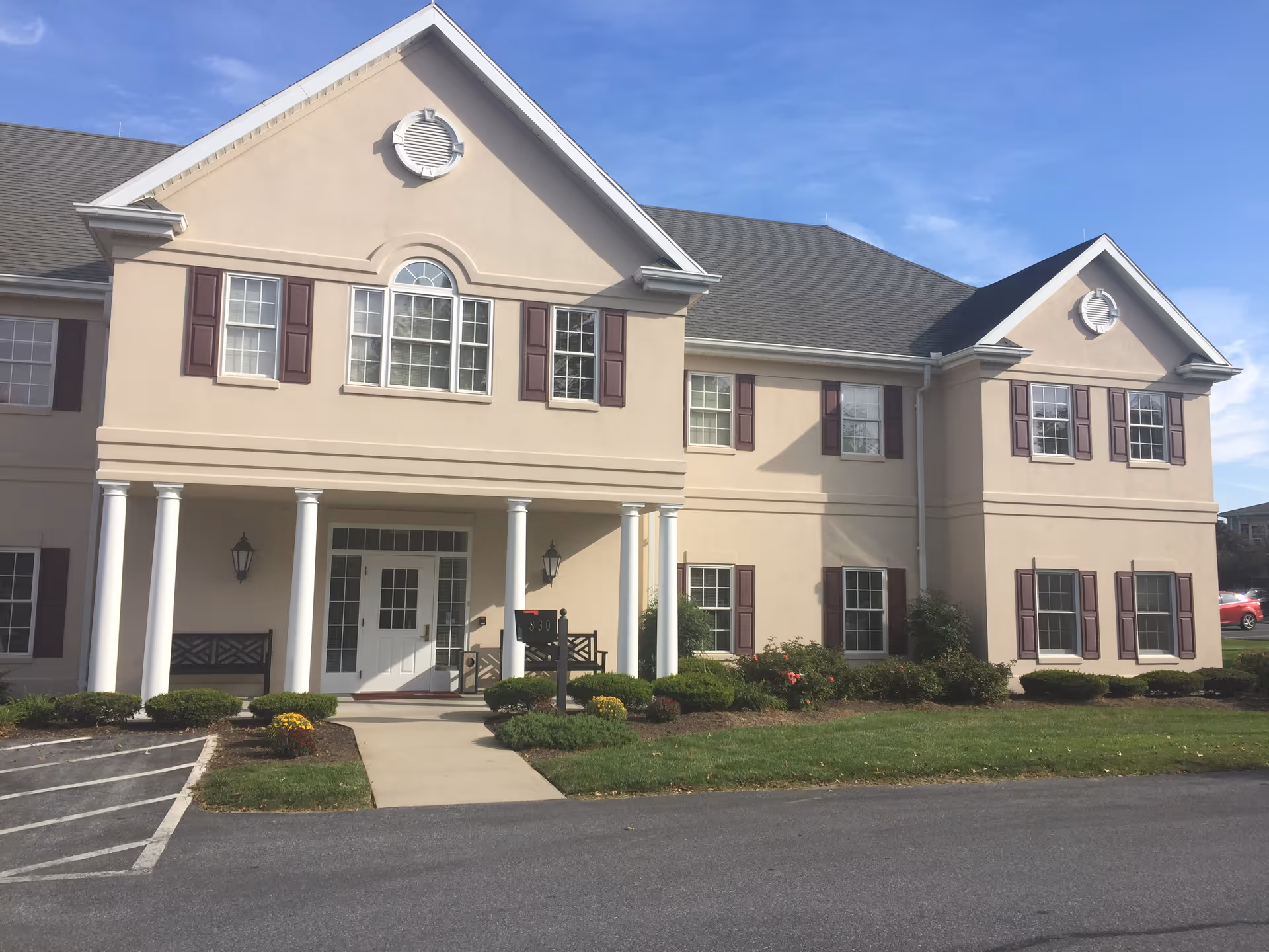 Exterior view of a two-story beige building with white columns at the entrance, multiple windows with brown shutters, a small landscaped garden with bushes and flowers, and a paved driveway in front under a blue sky.