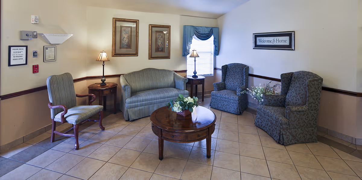 Cozy senior living facility sitting area with a sofa, armchairs, coffee table, lamps, and framed wall art.