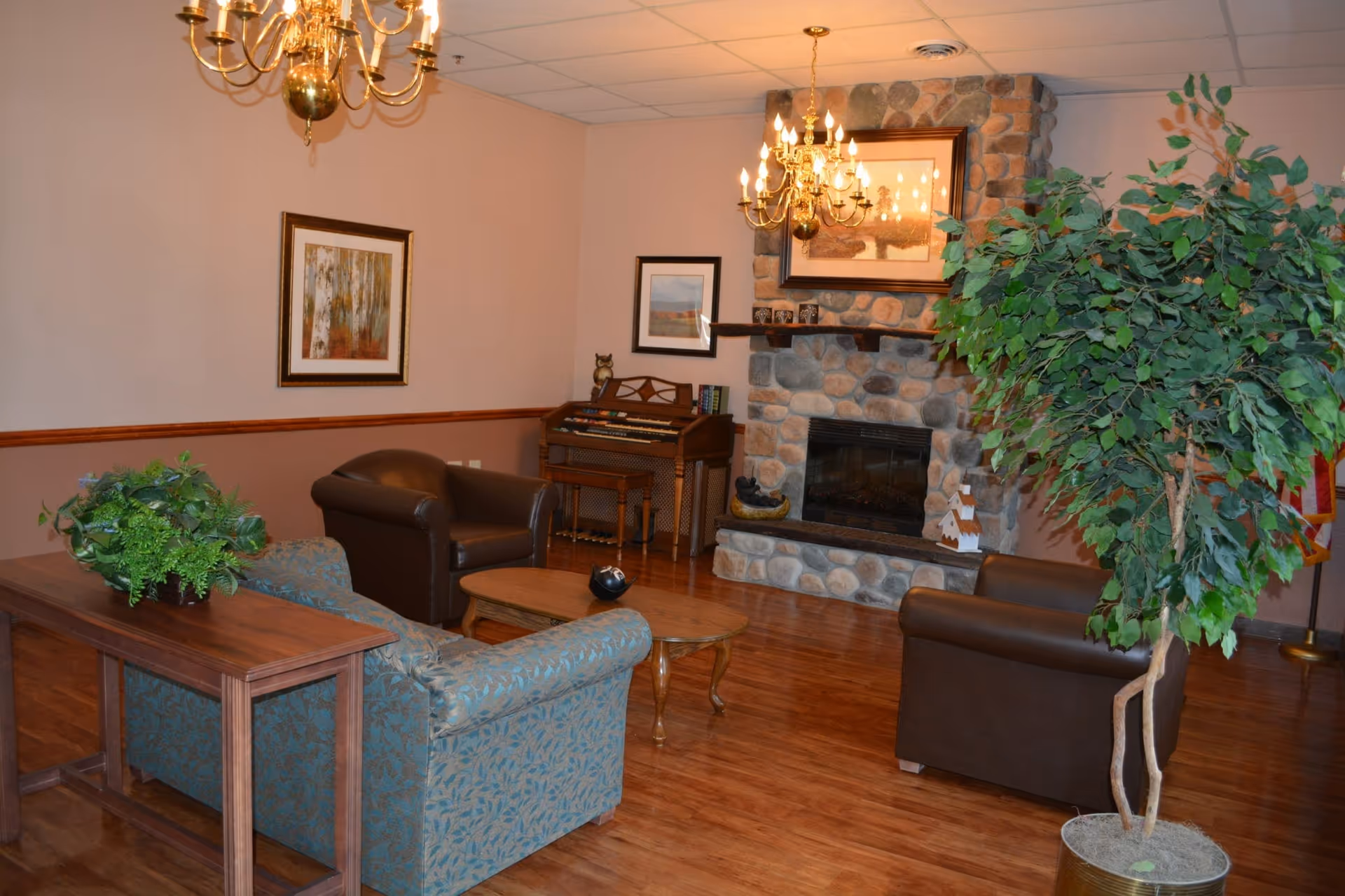 A cozy living room area with wooden flooring, a stone fireplace with a framed picture above it, two brown leather armchairs, a blue patterned sofa, a wooden coffee table, a wooden console table with a green plant, a large potted plant, two chandeliers, and a small organ against the wall with framed pictures above it.
