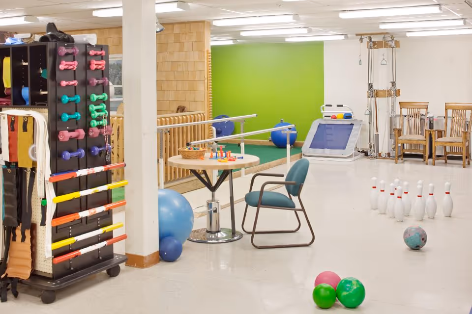 A rehabilitation room with various physical therapy equipment including colorful dumbbells, resistance bands, exercise balls, a table with therapy tools, a chair, and a bowling setup with pins and balls. The room has a green accent wall and wooden chairs along the back wall.
