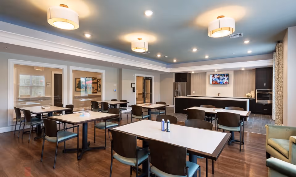 A spacious dining room with multiple tables and chairs arranged neatly on a wooden floor. The room features modern ceiling lights, a kitchenette area with a refrigerator, oven, and sink, and a wall-mounted television. Large windows and glass doors provide natural light, and there is a comfortable armchair in the corner.