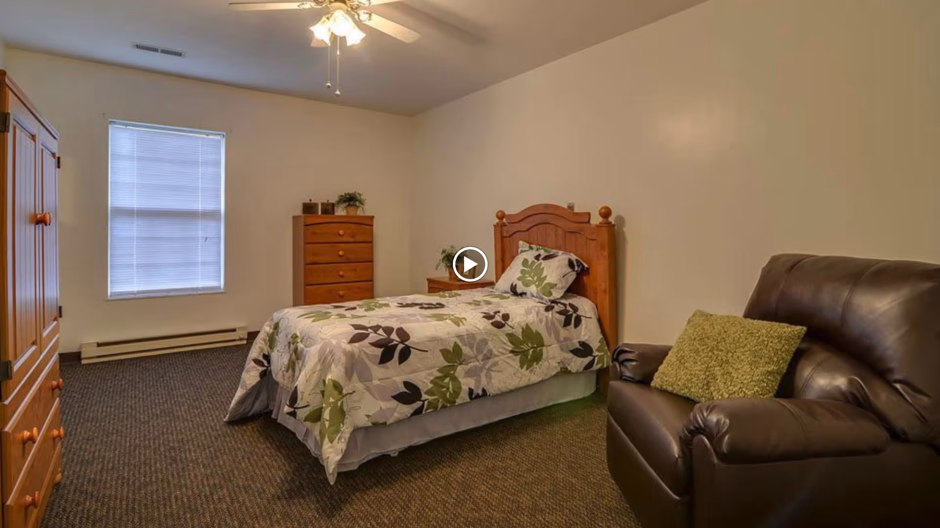 Simple bedroom with a single bed dressed in floral bedding, wooden headboard and dressers, a leather recliner with a green pillow, and a window with blinds.