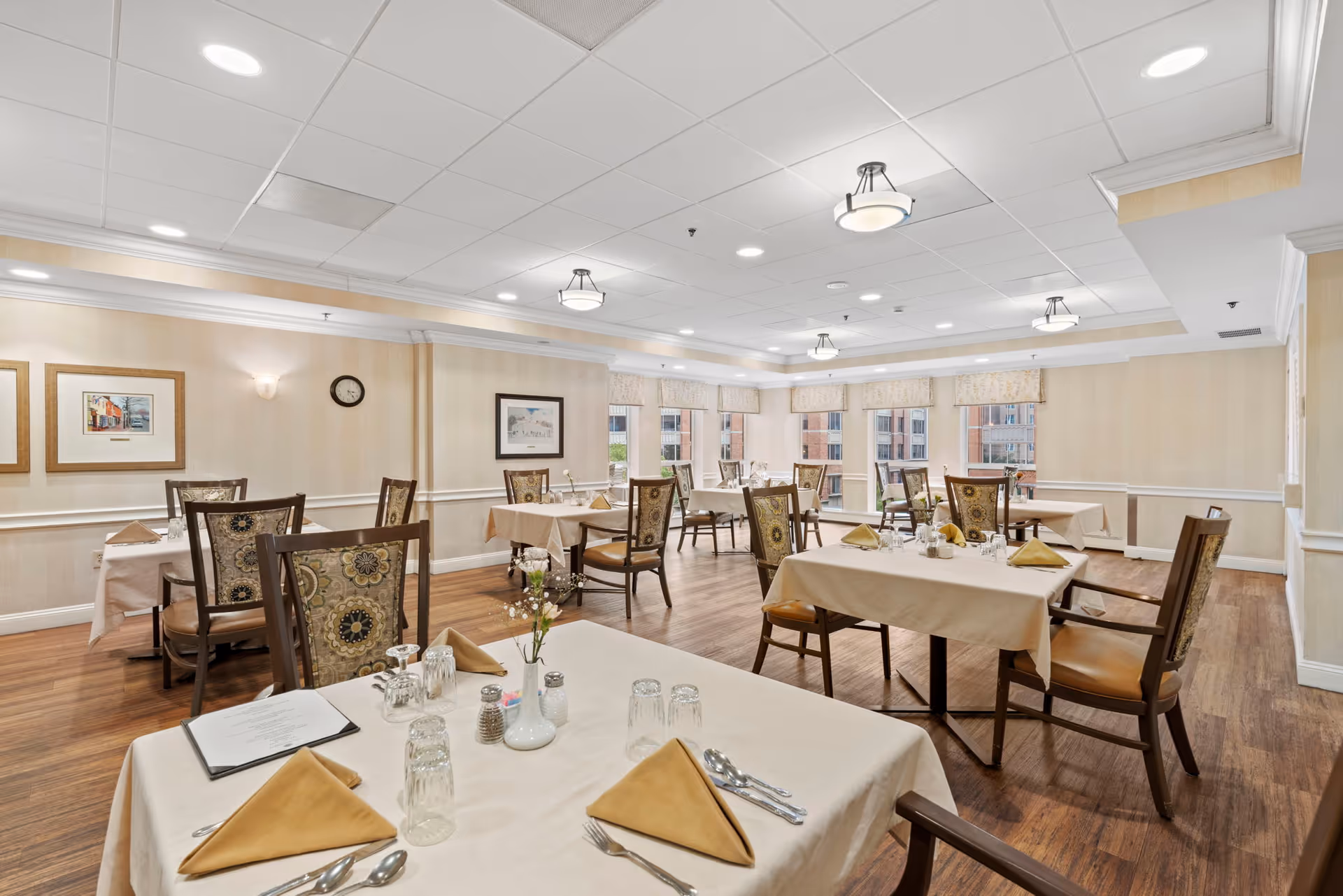 A bright and spacious dining room with multiple tables covered in beige tablecloths, each set with folded napkins, glasses, silverware, and small flower vases. The room has wooden flooring, cream-colored walls adorned with framed artwork, and large windows letting in natural light. Ceiling lights provide additional illumination.
