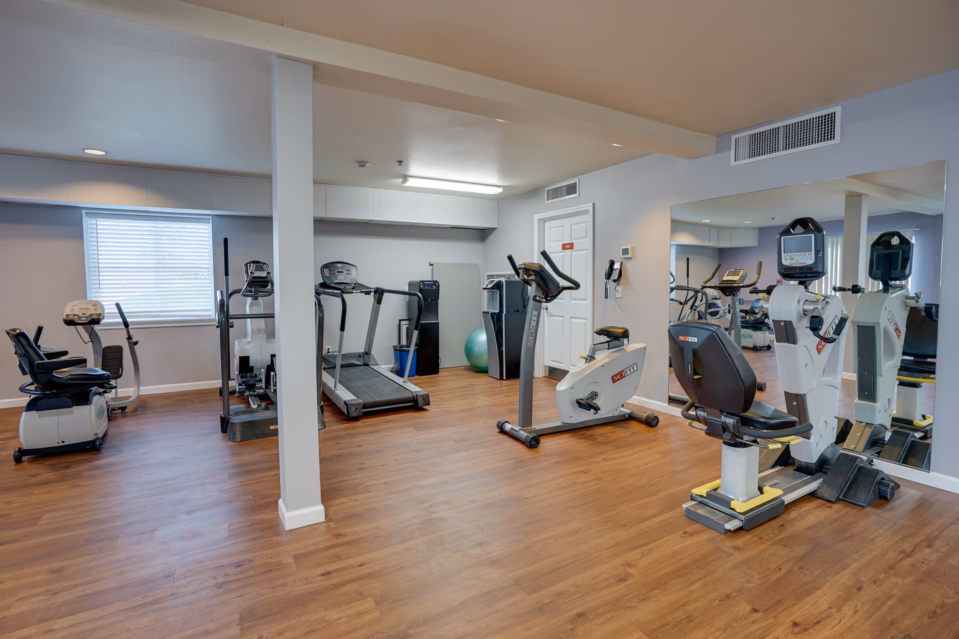 Well-lit fitness room with treadmills, stationary bikes, and exercise equipment on wood flooring and a large mirror.