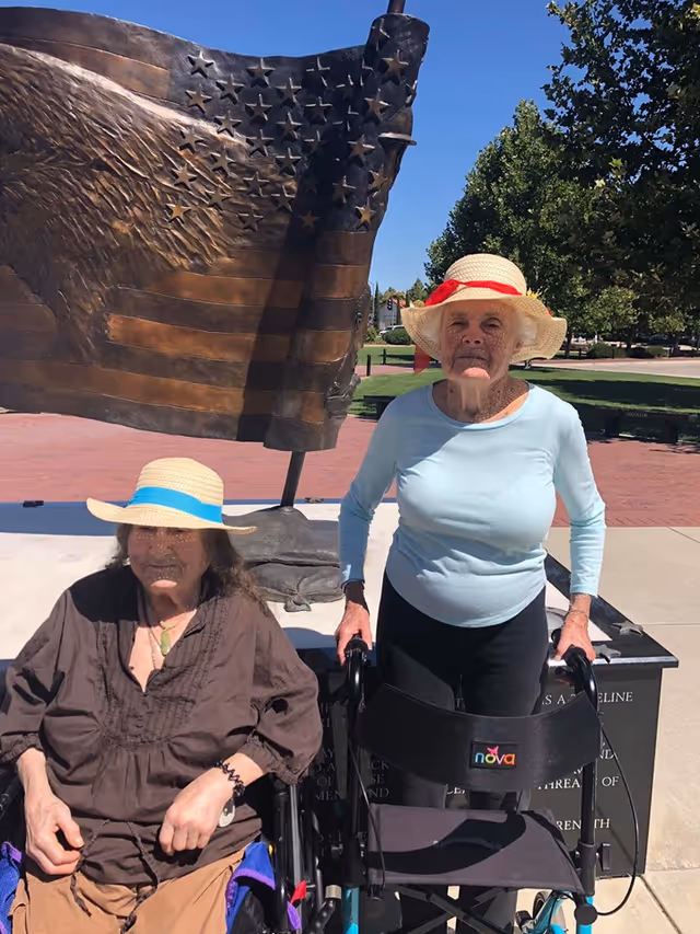 Two elderly women outdoors in front of a large bronze sculpture of an American flag. One woman is sitting in a wheelchair wearing a brown top and a straw hat with a blue band. The other woman is standing with the aid of a walker, wearing a light blue long-sleeve shirt and a straw hat with a red band. Trees and a clear blue sky are visible in the background.
