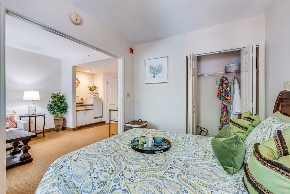 Interior view of a senior living facility room showing a bed with patterned bedding and green pillows in the foreground. An open closet with clothes hanging is visible on the right. The room opens into a small kitchenette area with cabinets, a refrigerator, and a plant. A living area with a lamp, side table, and armchair is partially visible on the left.