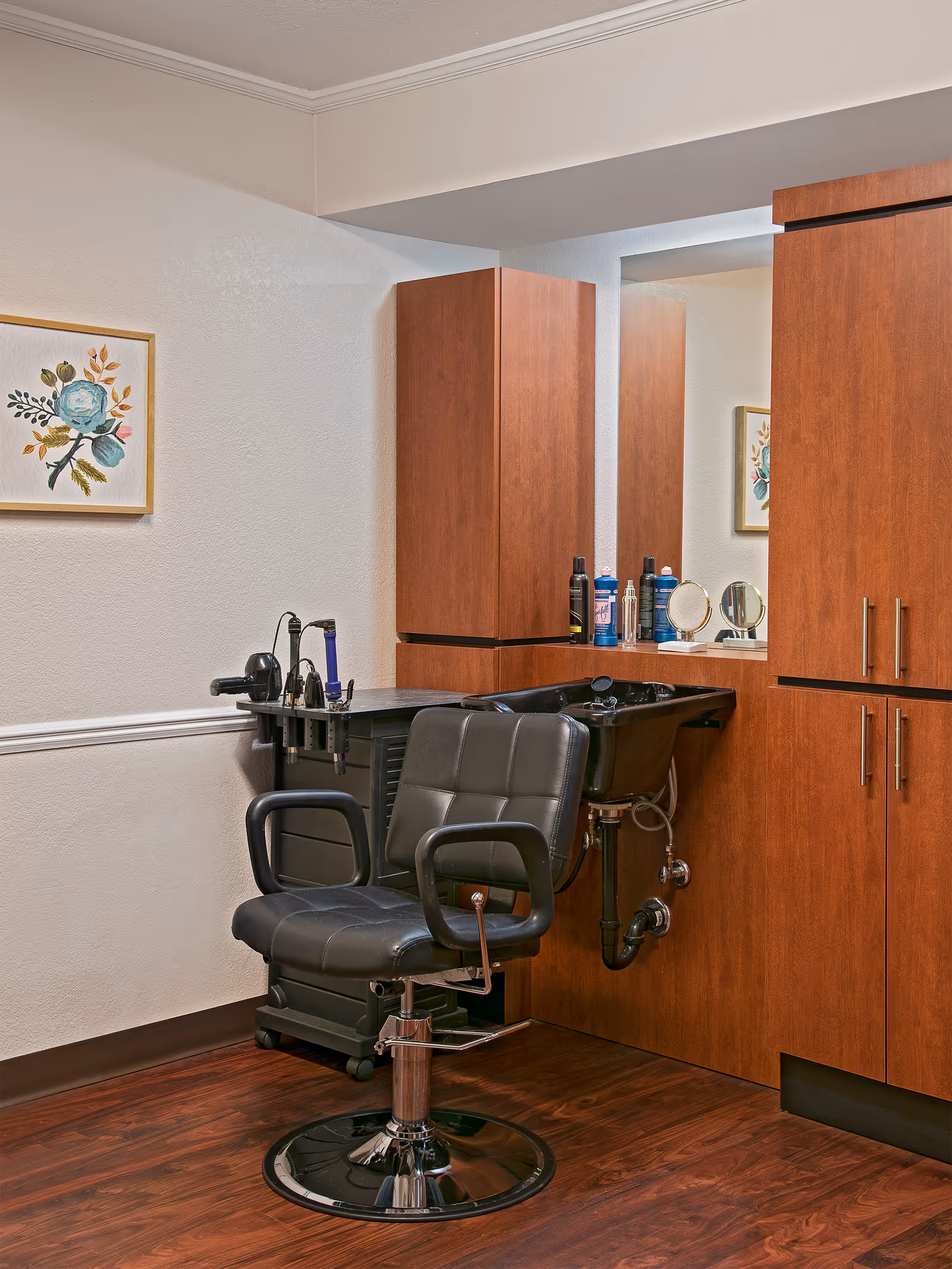 Interior view of a salon area in a senior living facility featuring a black salon chair in front of a black hair washing sink. The area has wooden cabinets, a mirror, and various hair care products on the countertop. A framed floral artwork is hung on the wall.