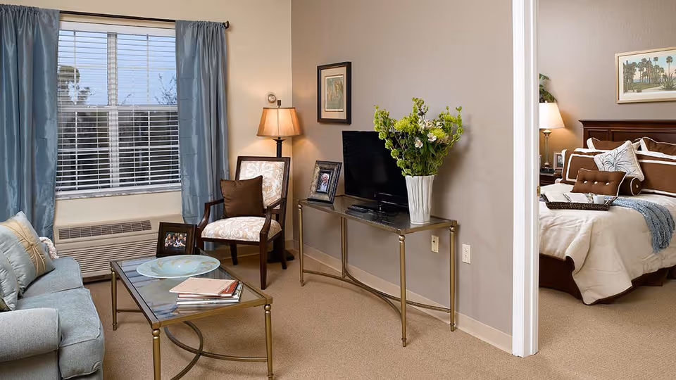 A cozy living room with a gray sofa, glass coffee table with books and a decorative plate, an armchair with a brown cushion, a floor lamp, and a TV on a slim console table with a vase of flowers. Through an open doorway, a bedroom is visible with a neatly made bed, decorative pillows, a bedside lamp, and framed artwork on the wall.