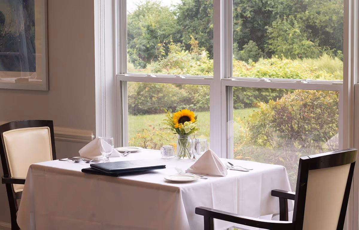 A dining table set for two with white tablecloth, napkins, plates, and silverware in front of a large window showing green bushes and trees outside. A vase with a sunflower and other flowers is placed in the center of the table.