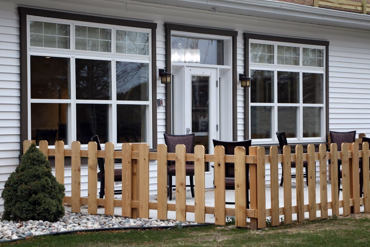 Outdoor patio area of a senior living facility with a wooden picket fence, several chairs, and large windows and a door leading inside the building.