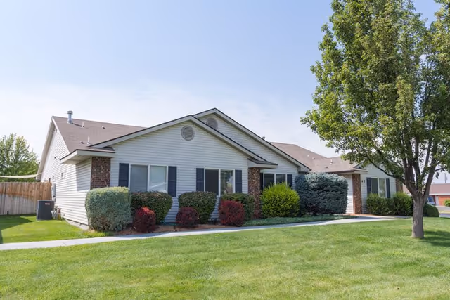 Single-story residential building with white siding and brick accents, surrounded by well-maintained green lawn, shrubs, and a large tree on the right side under a clear blue sky.