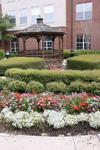 A wooden gazebo with a shingled roof is situated in a garden area surrounded by neatly trimmed green bushes and colorful flowers in front of a brick and beige building with multiple windows.