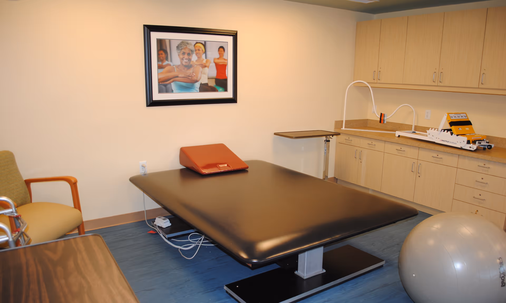 A therapy or rehabilitation room with a large black treatment table topped with a red wedge cushion, a silver exercise ball, a wooden chair with green upholstery, a small adjustable table, and light wood cabinets along the wall. A framed picture of smiling people exercising hangs on the beige wall.