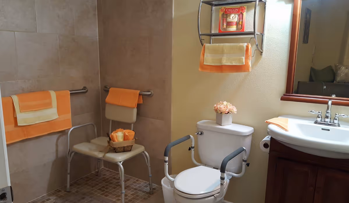 Accessible bathroom with a shower area featuring grab bars and a shower chair. The toilet has arm supports for assistance, and there is a sink with a mirror above it. Orange and beige towels are neatly placed on the towel racks, shower chair, and sink. A small flower arrangement is on top of the toilet tank.