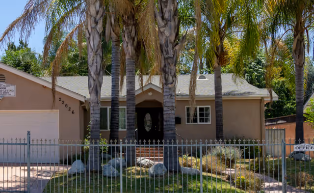 Single-story beige senior living facility front with palm trees, iron fence, and a gated yard.