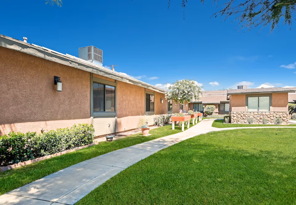 Courtyard with a paved walkway, green lawn, planter boxes and single-story stucco buildings under a blue sky.