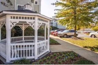 White wooden gazebo on landscaped grounds beside a sidewalk with parked cars and a multi-story building in the background.