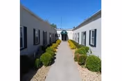 Outdoor walkway between two single-story white buildings with black window shutters and green bushes lining the path under a clear blue sky.