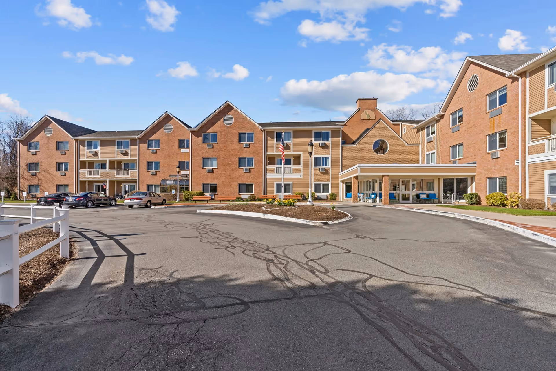 Exterior view of a senior living facility with a large brick building featuring multiple windows and balconies. There is a circular driveway with several parked cars, an American flag on a pole, and a covered entrance area with seating. The sky is blue with scattered clouds.