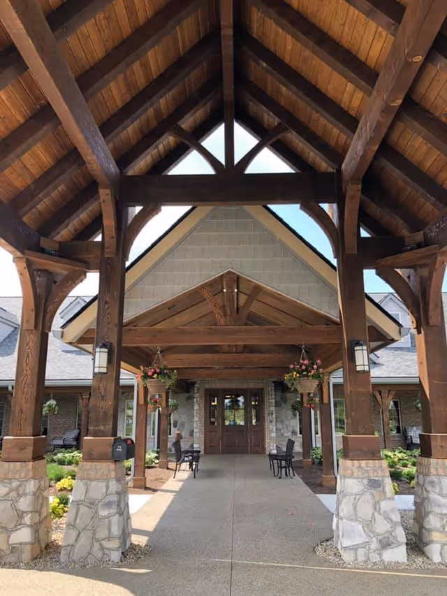 Entrance of The Inn at Library Way featuring a covered walkway with wooden beams and stone pillars, hanging flower baskets, outdoor chairs, and a double wooden door under a peaked roof.