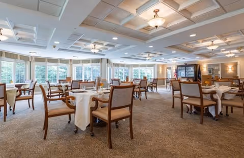 Spacious dining room with multiple tables covered with white tablecloths and wooden chairs arranged around them. Large windows with curtains allow natural light to fill the room. The ceiling has decorative panels and several light fixtures. The room has a carpeted floor and an American flag is visible near a cabinet in the background.