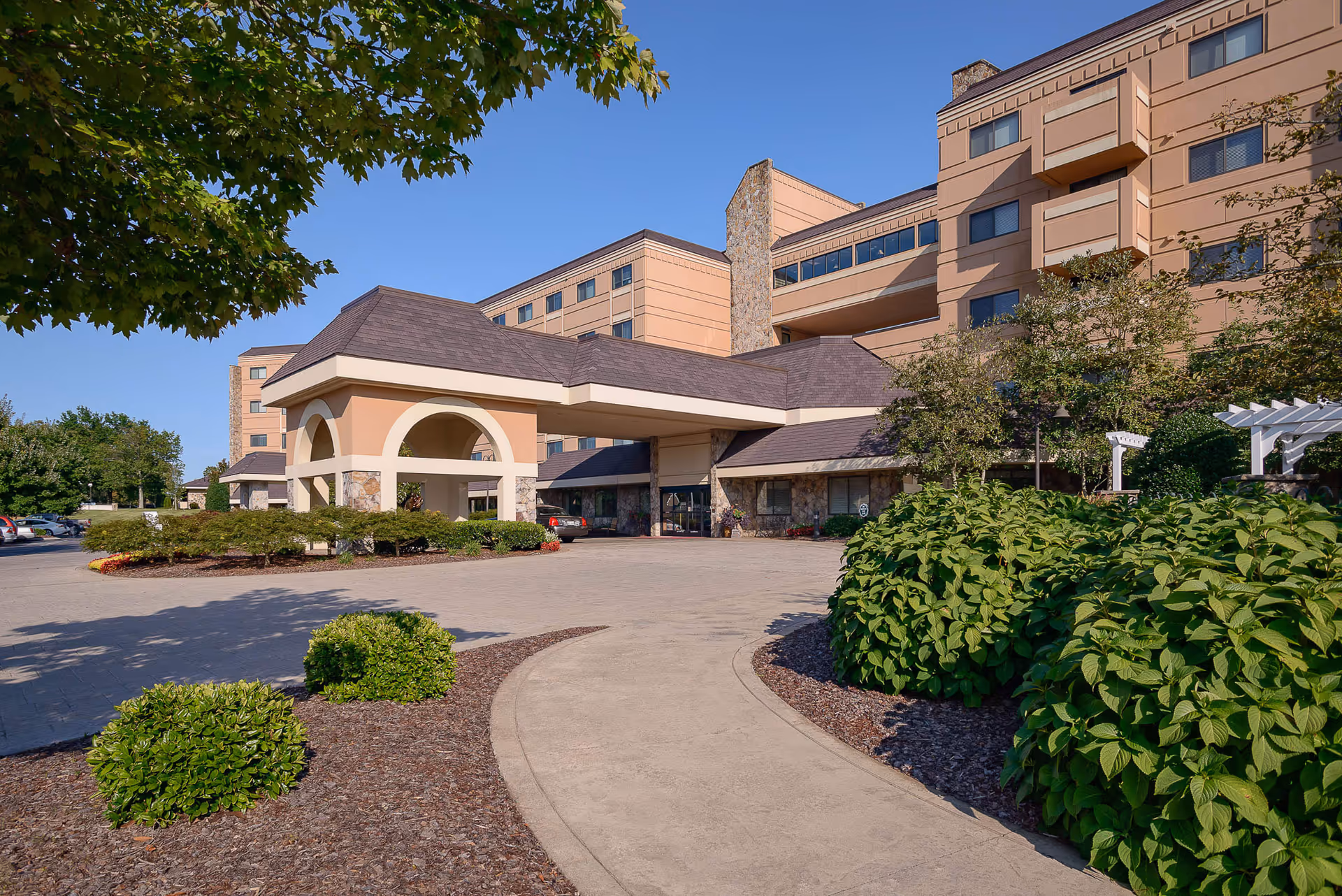 Front entrance and porte-cochère of a multi-story senior living facility with landscaped driveway and shrubs under a clear blue sky.
