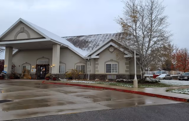 Front of a one-story residential building with a covered entrance, wet driveway, light snow on the roof and lawn, and a few parked cars.