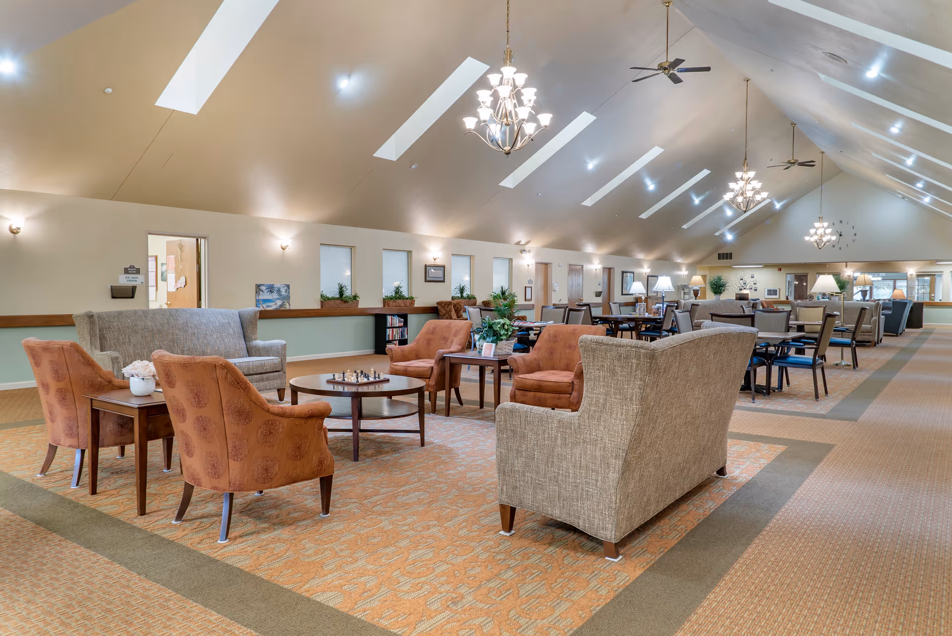 A spacious senior living facility common area with high vaulted ceilings and skylights. The room features multiple seating arrangements including beige and orange upholstered armchairs and sofas around wooden coffee tables. There are ceiling fans, chandeliers, and several lamps providing warm lighting. The walls have small windows with plants on the ledges and various framed pictures. In the background, there are more tables and chairs arranged for dining or activities.