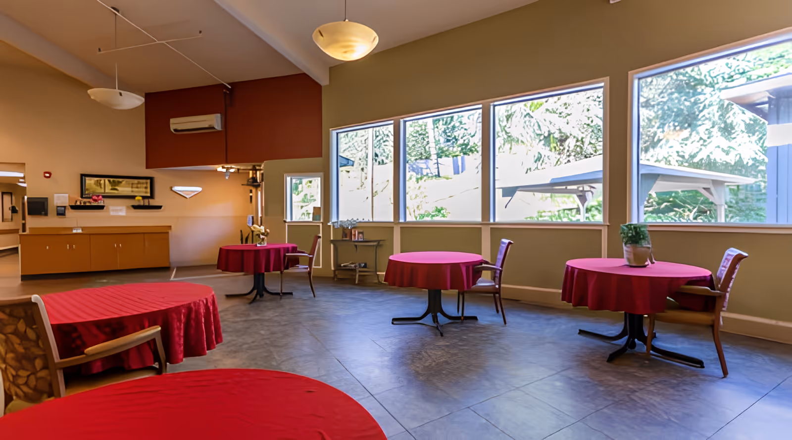 Bright communal dining area with round tables covered in red tablecloths, chairs, and large windows overlooking greenery.