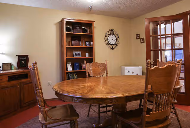 A dining room with a round wooden table surrounded by chairs, a bookshelf, wall clock, and French doors.