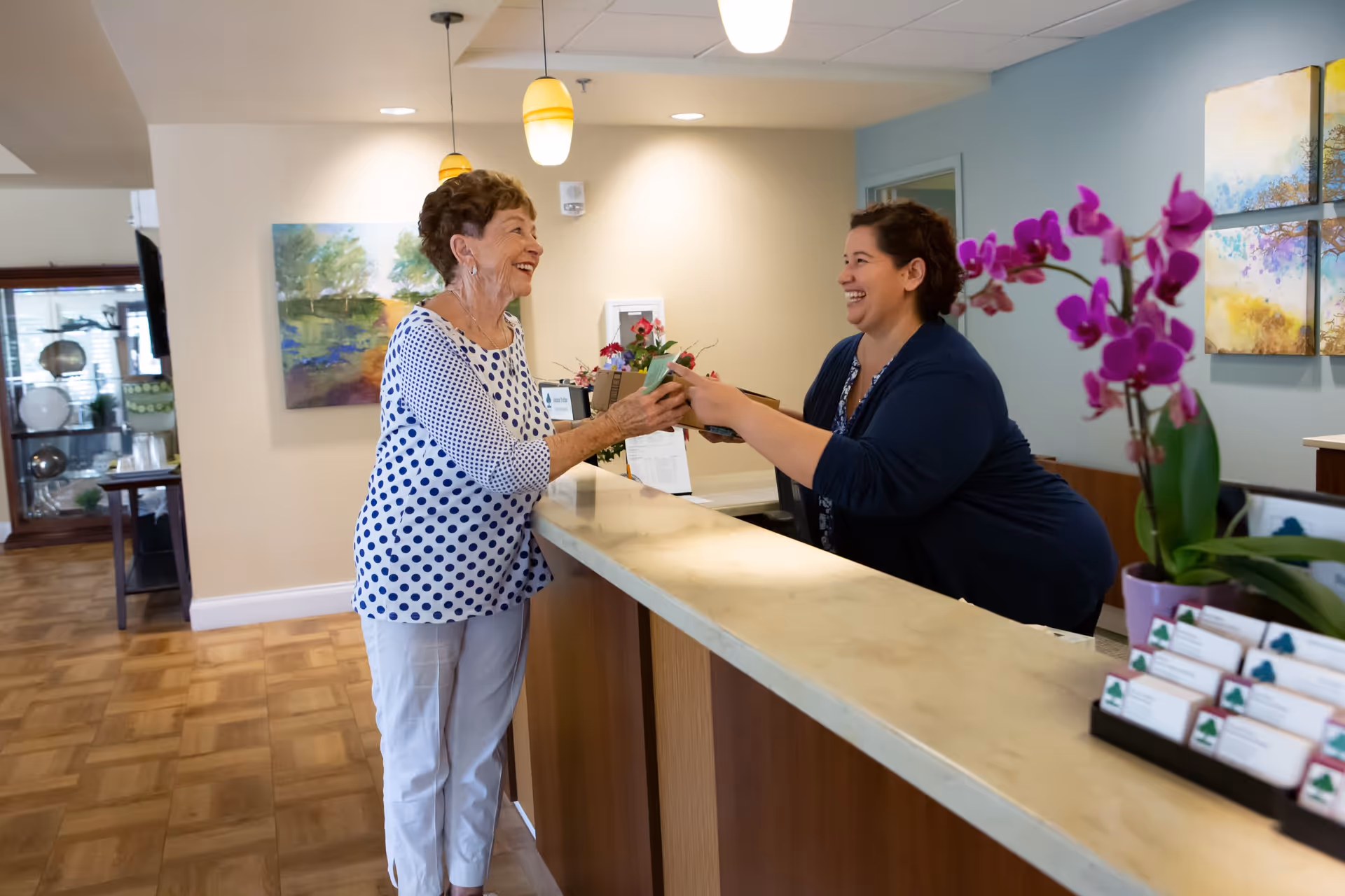 An elderly woman in a white and blue polka dot shirt is smiling and receiving a small box from a smiling receptionist behind a counter in a well-lit interior space with paintings and flowers.