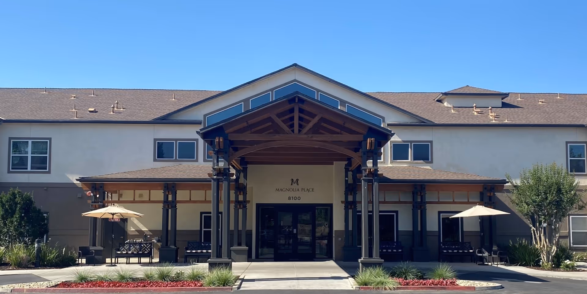 Front exterior view of Magnolia Place facility with a covered entrance supported by dark pillars, seating areas with umbrellas on both sides, and a clear blue sky above.